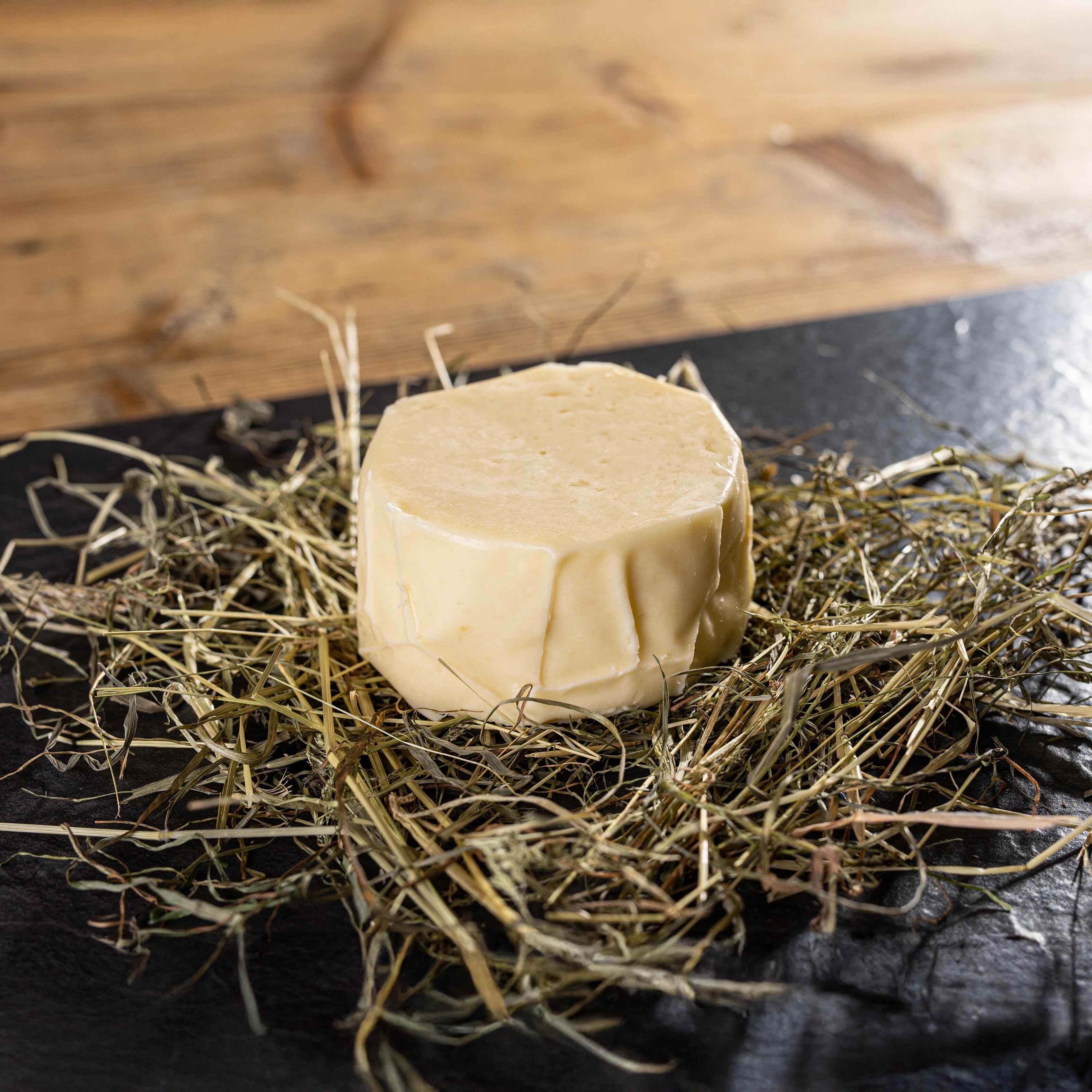 Ein runder Alpbachtaler Mugl-Käse liegt auf einer Schicht Heu auf dunkler Platte.
