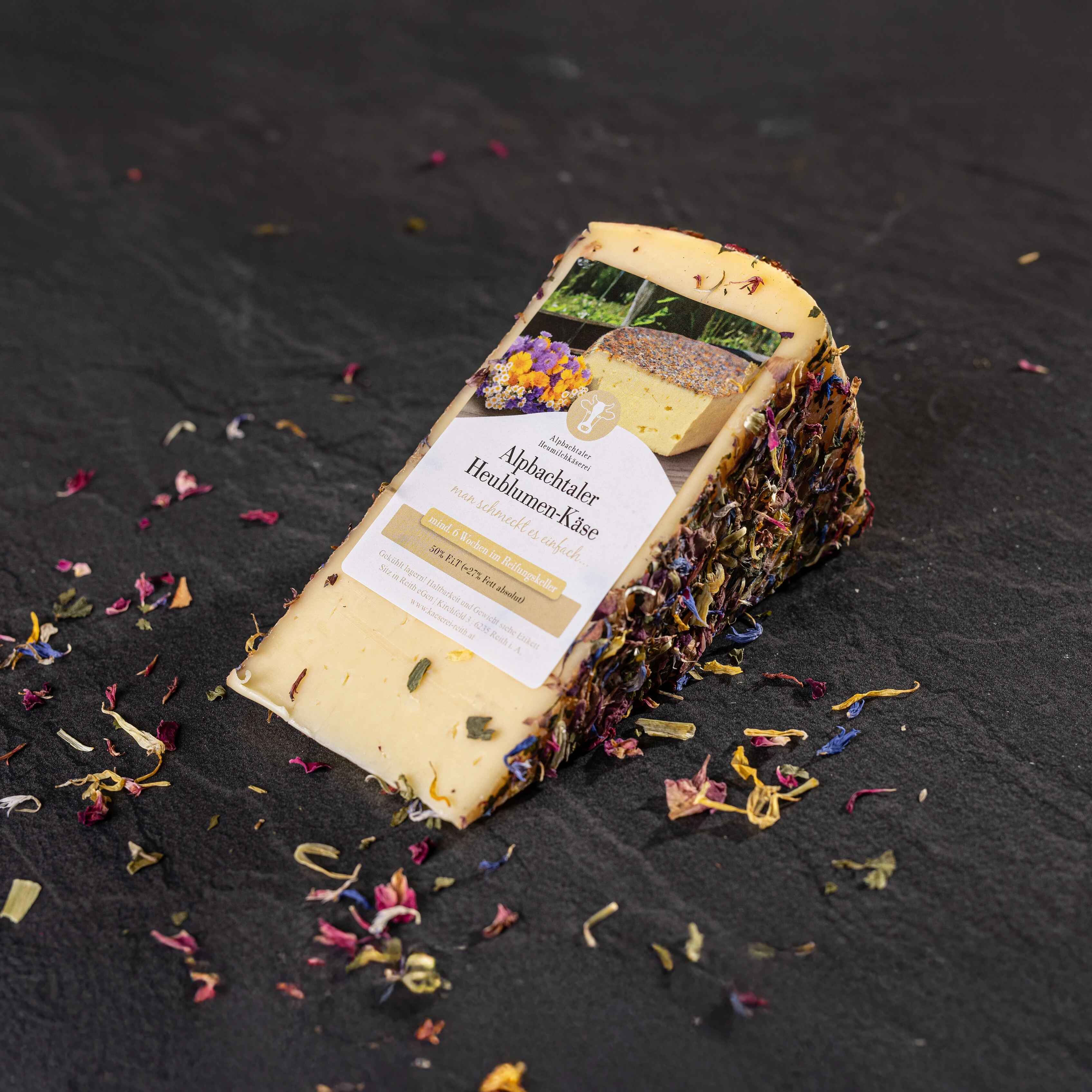 Ein Stück Alpbachtaler Heublumenkäse mit bunter Kräuter- und Blütenkruste liegt auf dunklem Steinuntergrund.