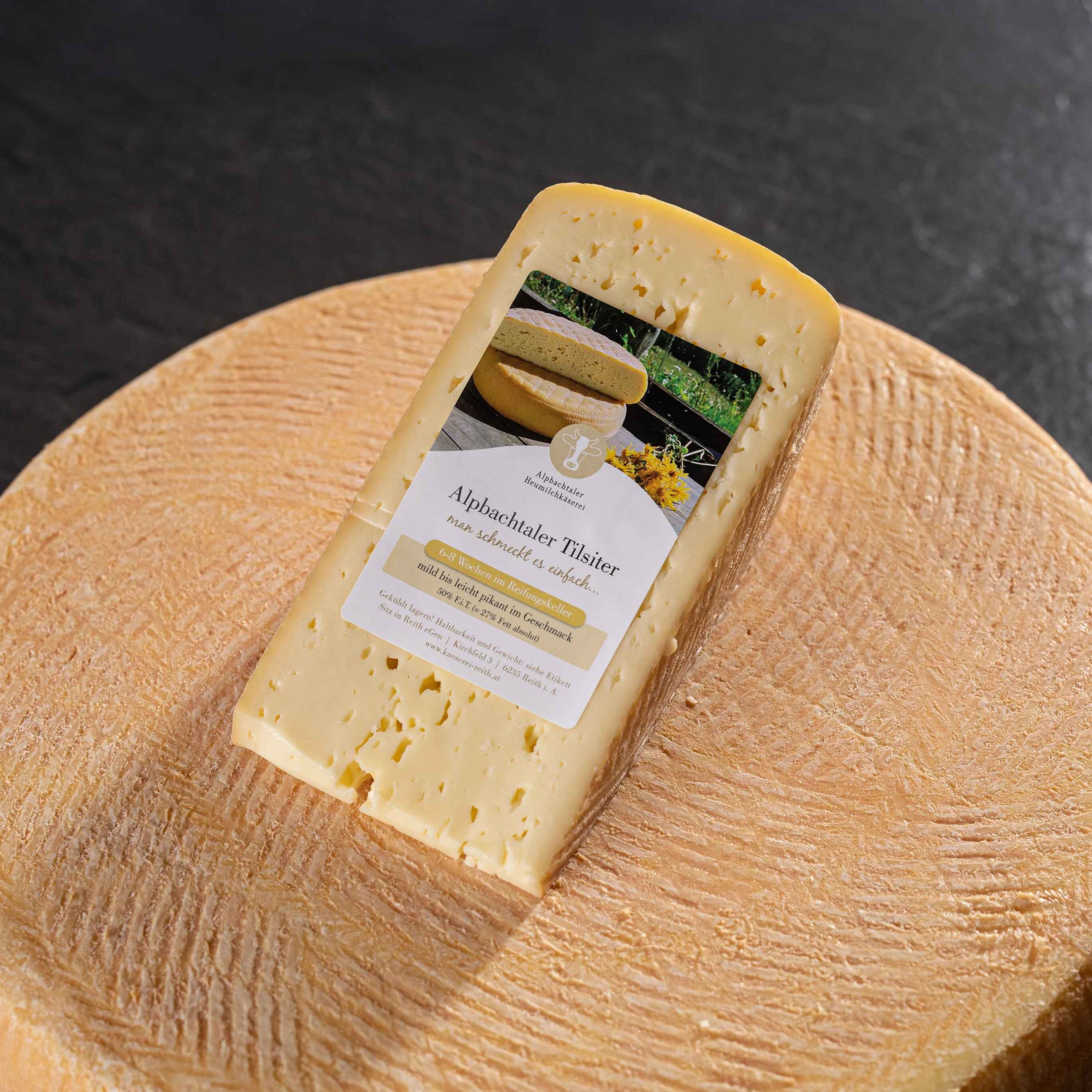 Alpbachtaler Tilsiter Ein Stück Alpbachtaler Tilsiter-Käse mit Etikett liegt auf einem großen runden Käselaib.