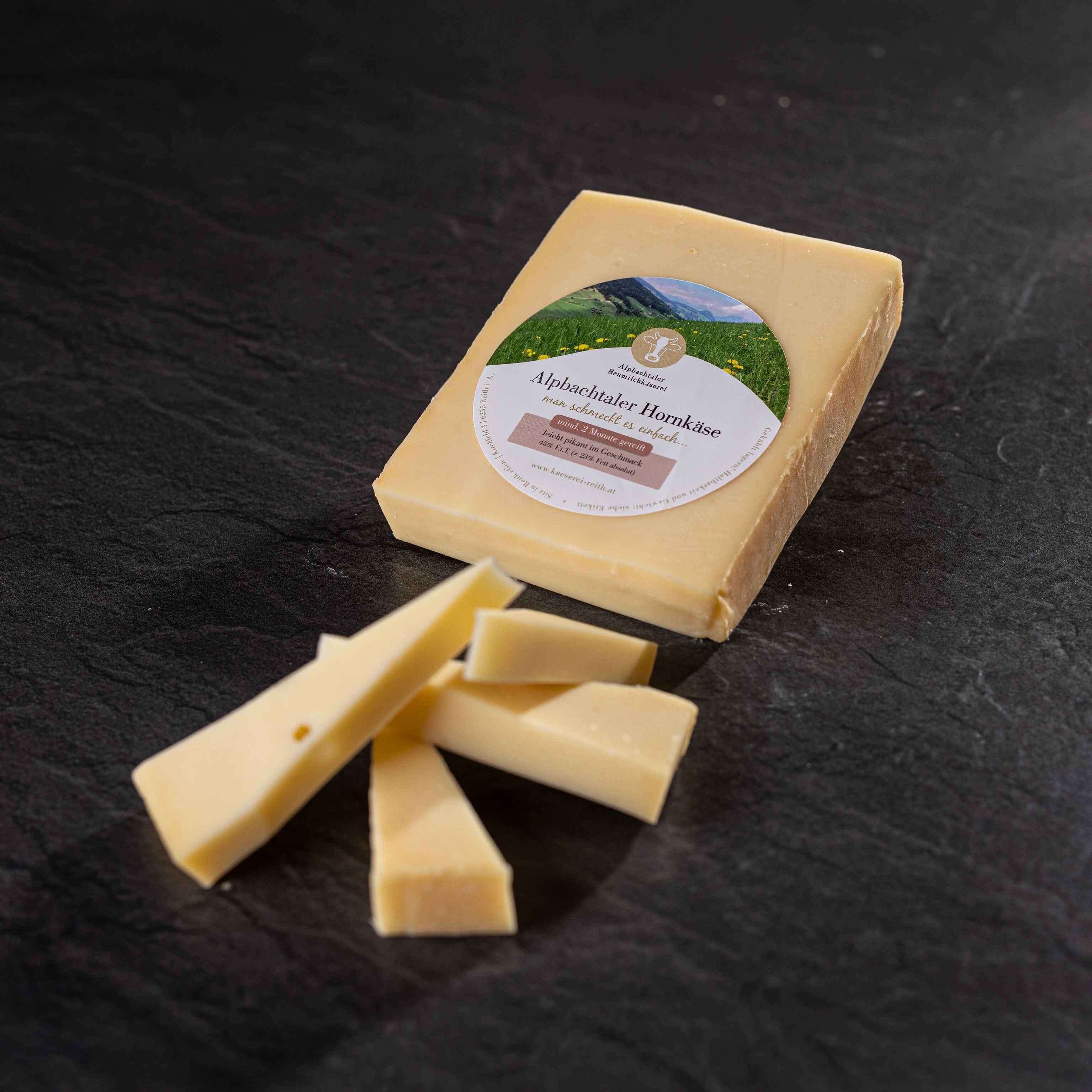 Ein Stück Alpbachtaler Hornkäse mit Etikett liegt auf dunklem Steinuntergrund, davor einige Käsestücke.