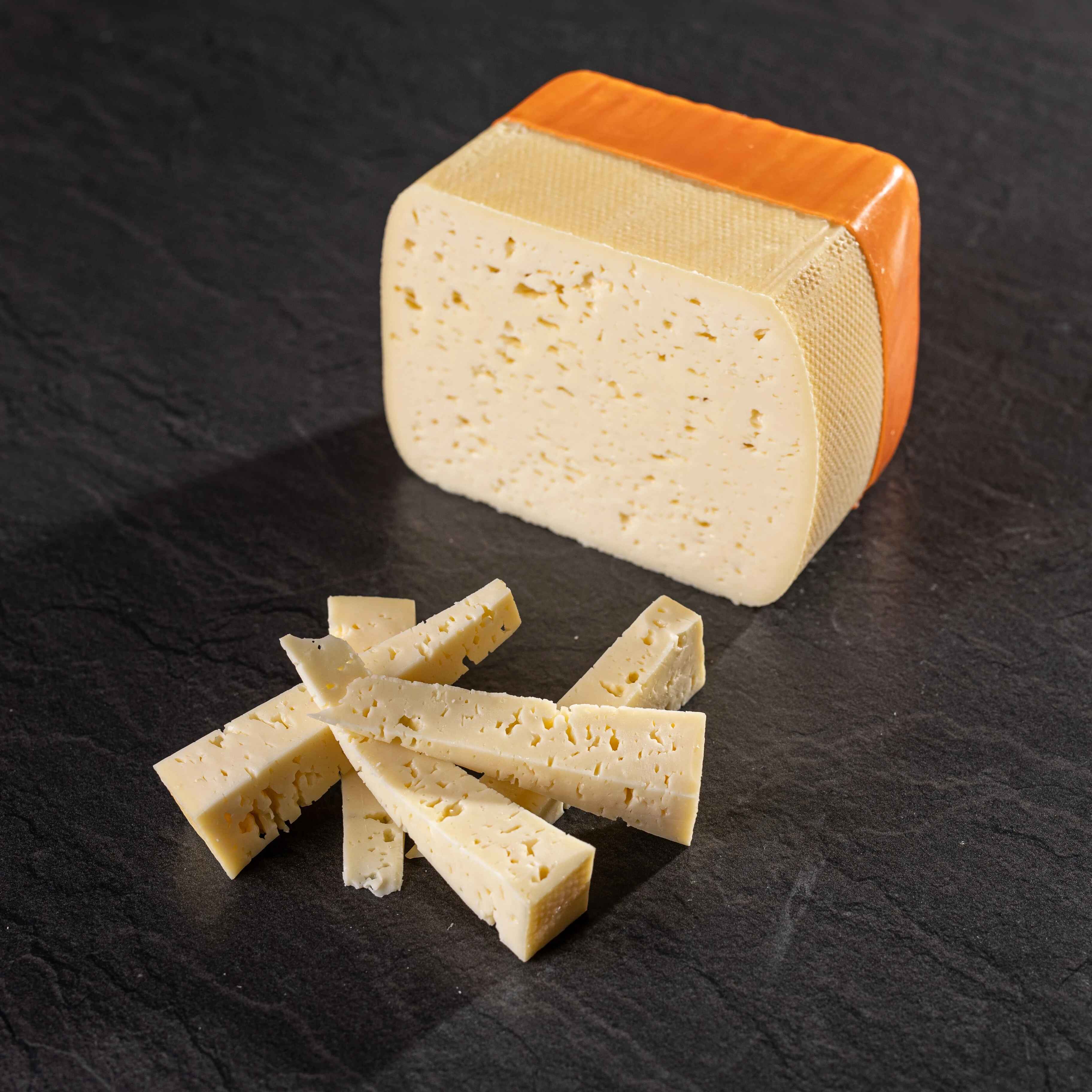 Alpbachtaler Kellergold Ein Stück Alpbachtaler Kellergold mit orangefarbener Rinde und einige geschnittene Käsesticks liegen auf dunklem Steinuntergrund.