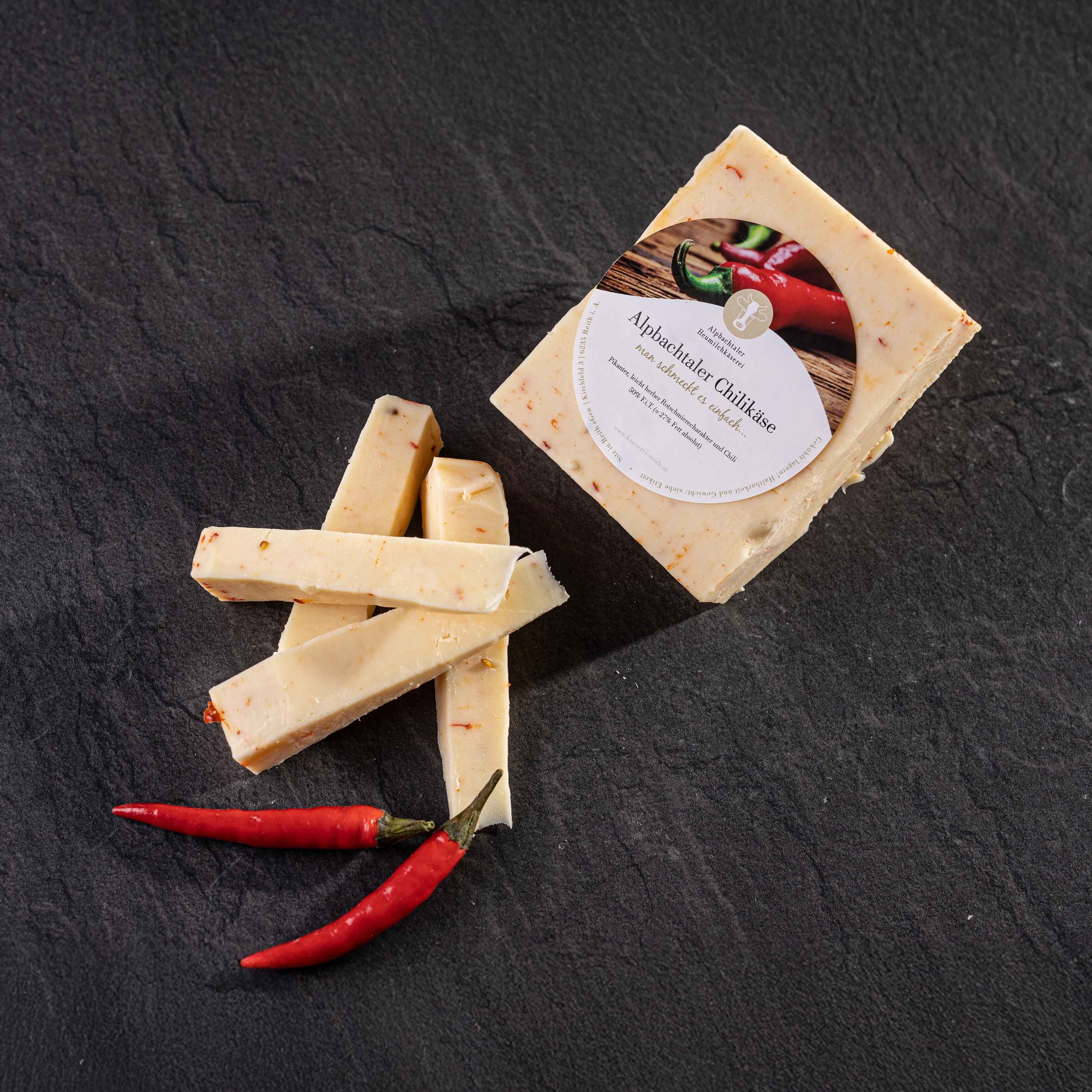 Alpbachtaler Chilikäse Ein Stück Alpbachtaler Chilikäse mit Etikett liegt auf dunklem Steinuntergrund, daneben Käsestreifen und rote Chilischoten.