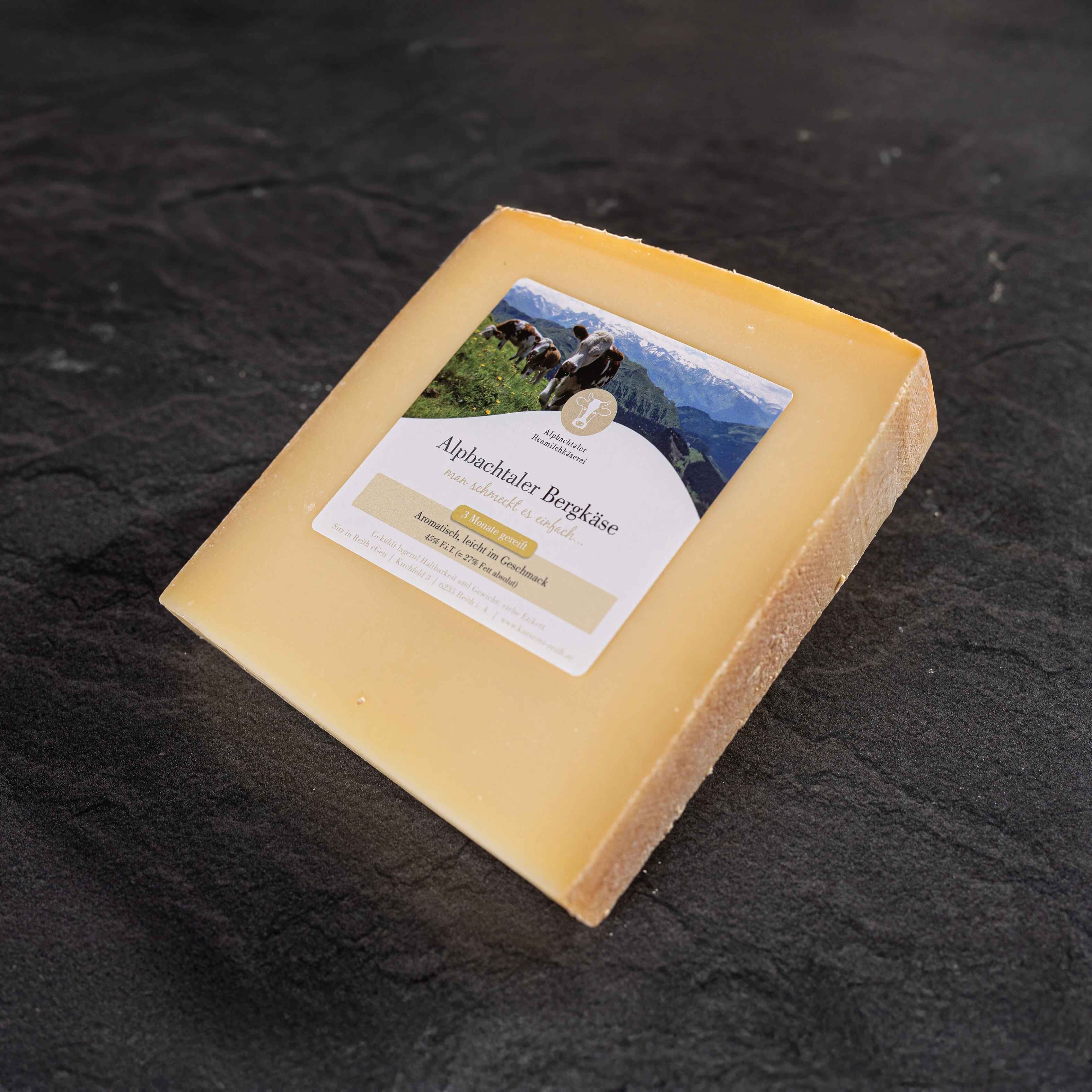 Ein Stück Alpbachtaler Bergkäse mit Etikett und heller Rinde liegt auf dunklem Steinuntergrund.