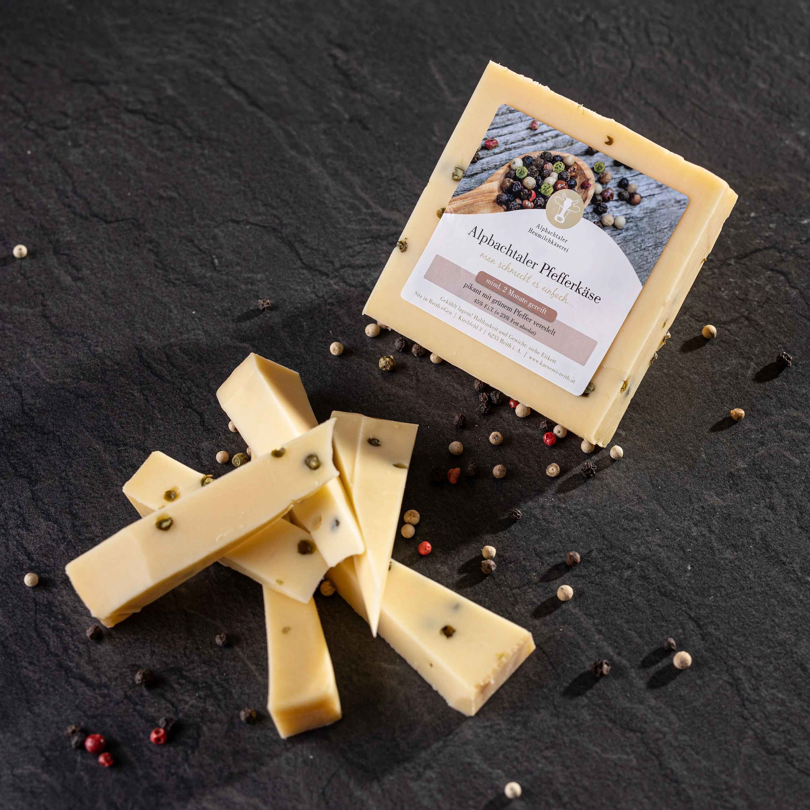 Alpbachtaler Pfefferkäse Ein Stück Alpbachtaler Pfefferkäse mit Etikett liegt auf dunklem Steinuntergrund, daneben Käsestreifen und bunte Pfefferkörner.