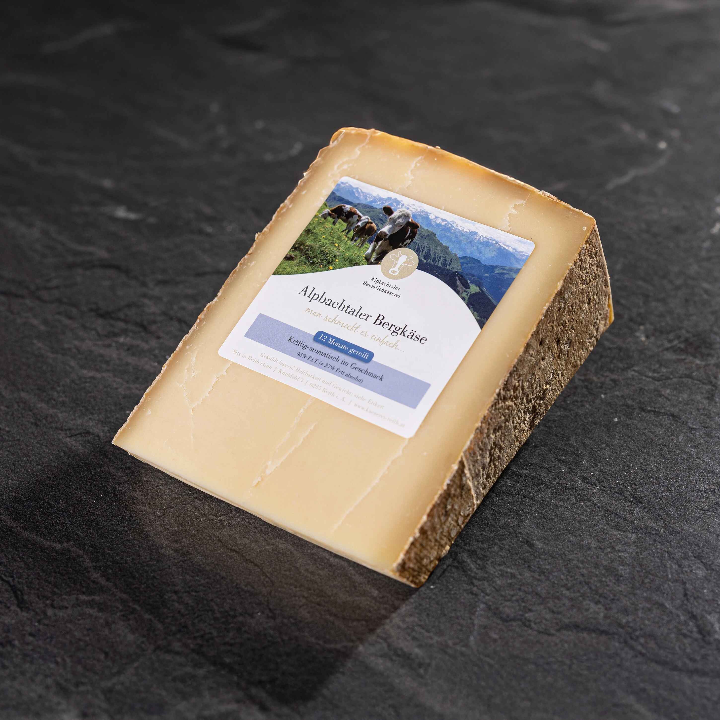 Alpbachtaler Bergkäse Ein Stück gereifter Alpbachtaler Bergkäse mit brauner Rinde und Etikett liegt auf dunklem Steinuntergrund.