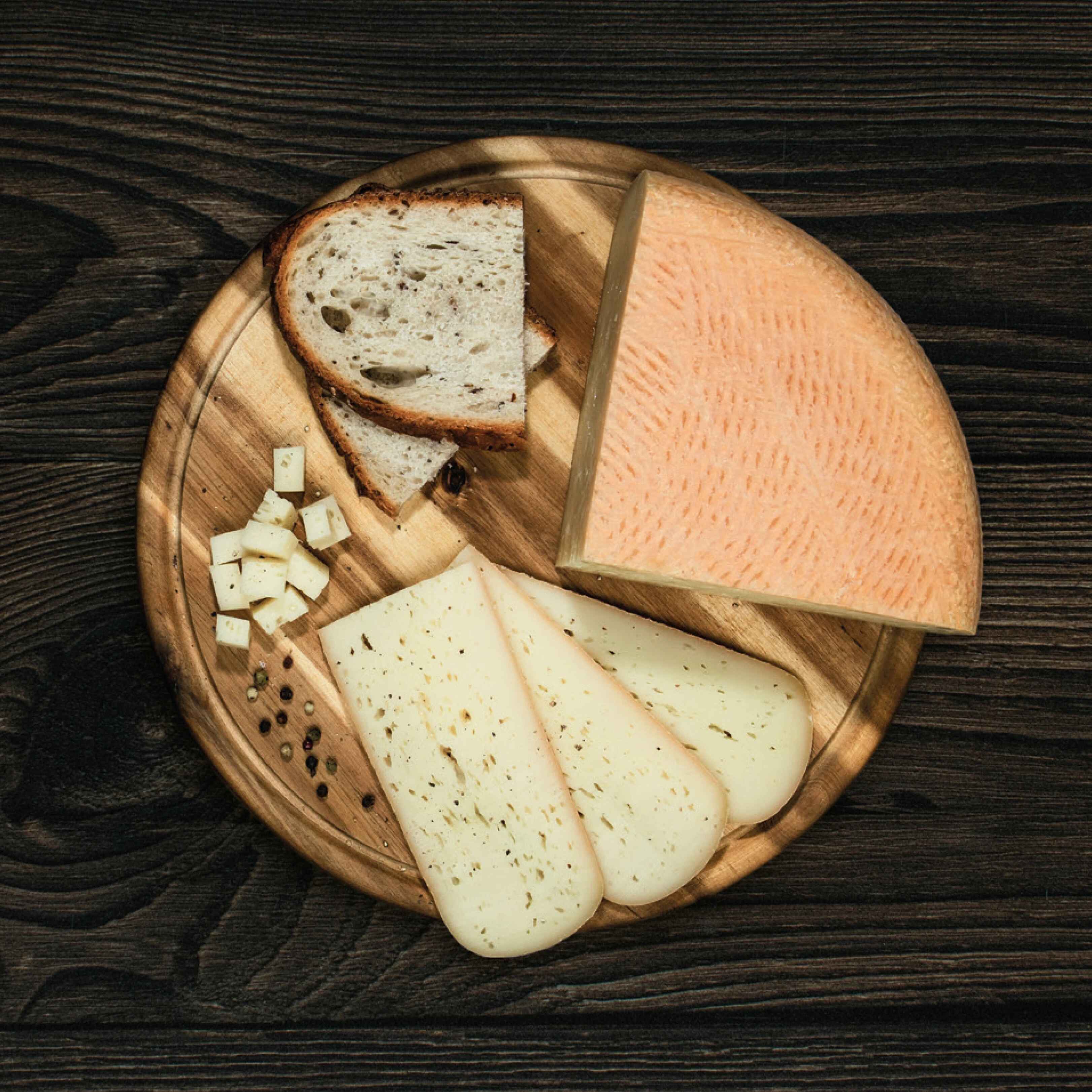 Alpbachtaler Bierkäse Ein Stück Alpbachtaler Bierkäse mit Brotscheiben, Käsescheiben und -würfeln liegt auf einem runden Holzbrett.